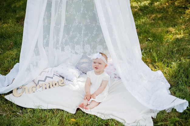 A little girl is sitting under a canopy on a white blanket in a Park. A cute girl looks away in a white dress and headband in nature.The little girl is naughty