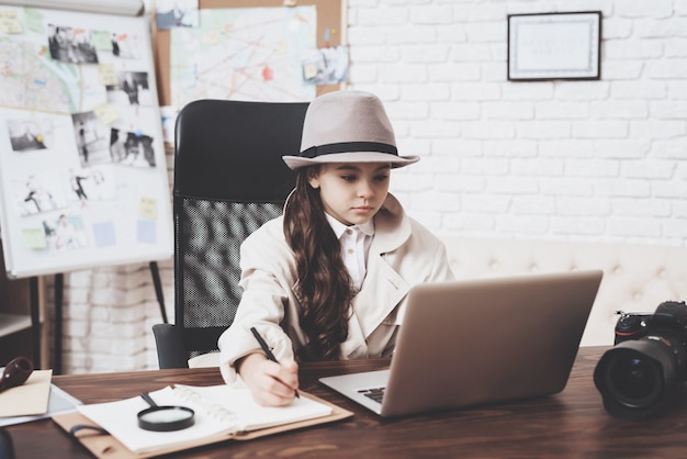 Little girl is sitting at desk taking notes near laptop.