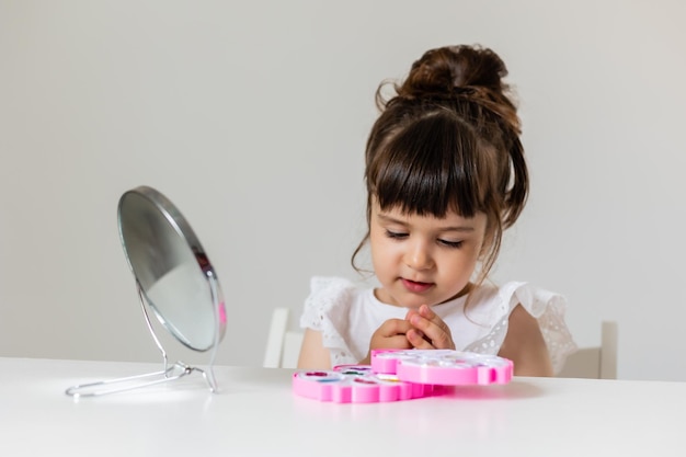 Little girl is sitting at the dressing table in front of the mirror and applying makeup like Mom