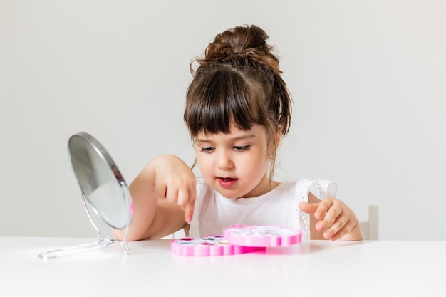 Little girl is sitting at the dressing table in front of the mirror and applying makeup like Mom