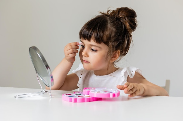 Little girl is sitting at the dressing table in front of the mirror and applying makeup like Mom