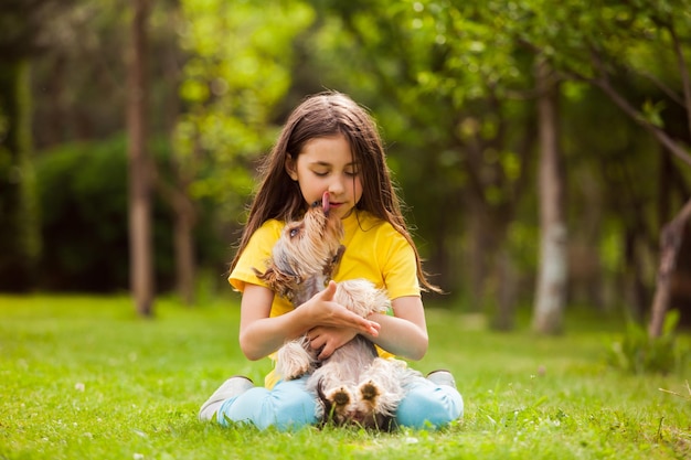 小さな女の子はヨークシャーテリアと一緒に緑の芝生に座っています犬は女の子をなめる