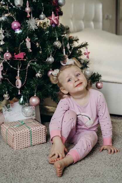 小さな女の子が贈り物を持ってクリスマスツリーで彼女のパジャマに座っています