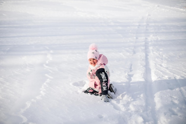 小さな女の子はピンクの冬のジャケットを着た雪の子供に座っており、ポンポン付きの帽子はnaで散歩を楽しんでいます...