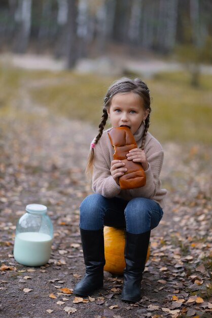 A little girl is sitting on a pumpkin, eating bread, next to a jar of milk