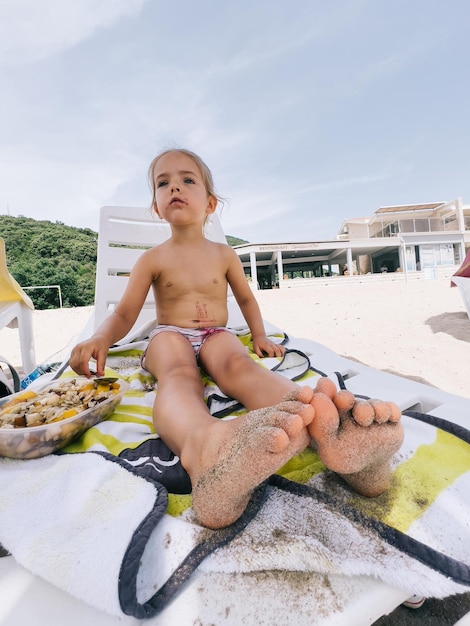 Little girl is sitting on a sun lounger on the beach next to a plastic box with food