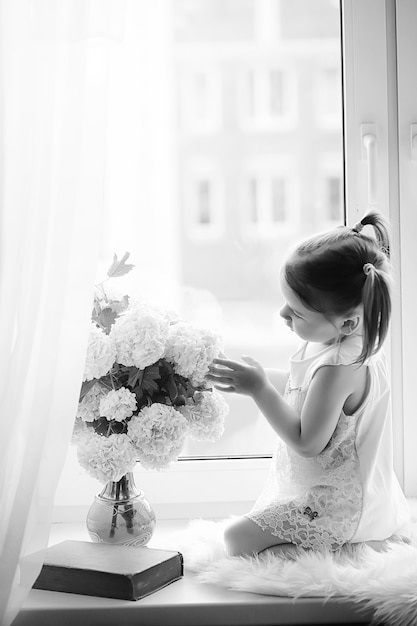 A little girl is sitting on the windowsill. A bouquet of flowers in a vase by the window and a girl sniffing flowers. A little princess in a white dress with a bouquet of white flowers by window.