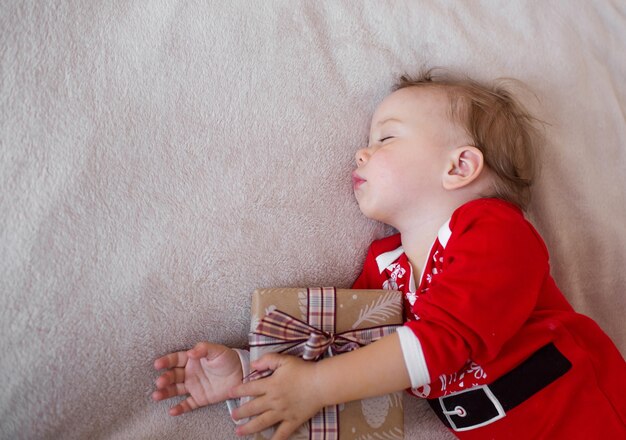 어린 소녀가 산타 클로스 의상을 입은 침대에서 자고 크리스마스 선물을 껴안고 있습니다.