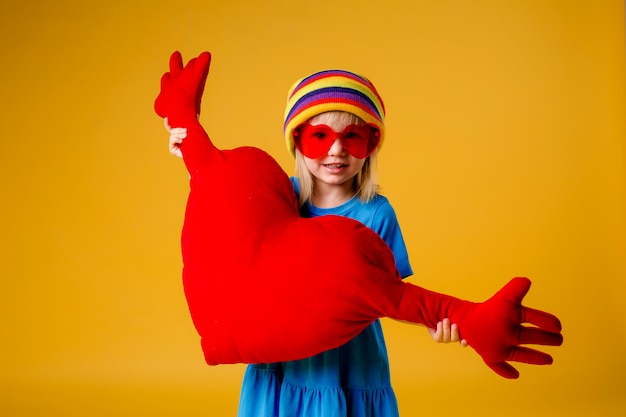 La bambina sorride tenendo un cuscino a forma di cuore rosso su uno sfondo giallo