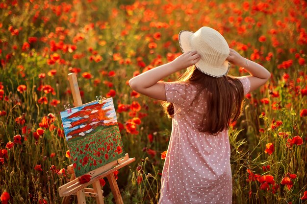 Little girl is standing in the field of red poppies, painted a landscape and admires the sunset