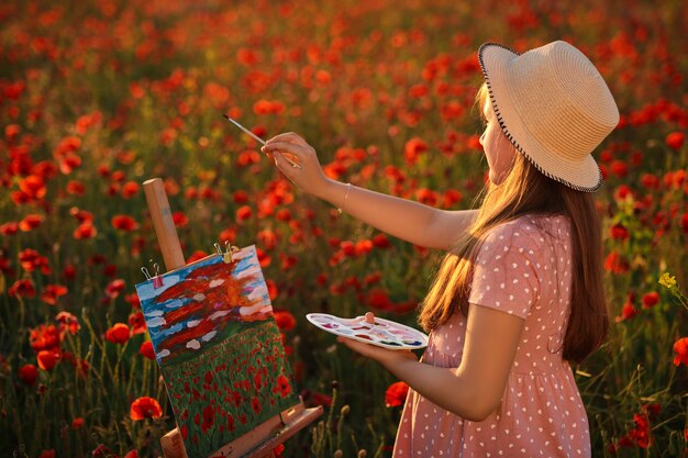 Little girl is standing in the field of red poppies On the Sunset and painting on the canvas placed on a drawing stand