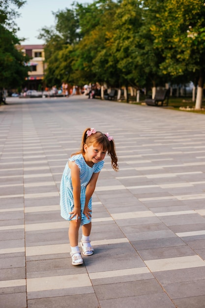 小さな女の子が公共の公園に立って、甘く微笑んでいます2つのポニーテールの美しいヘアスタイル