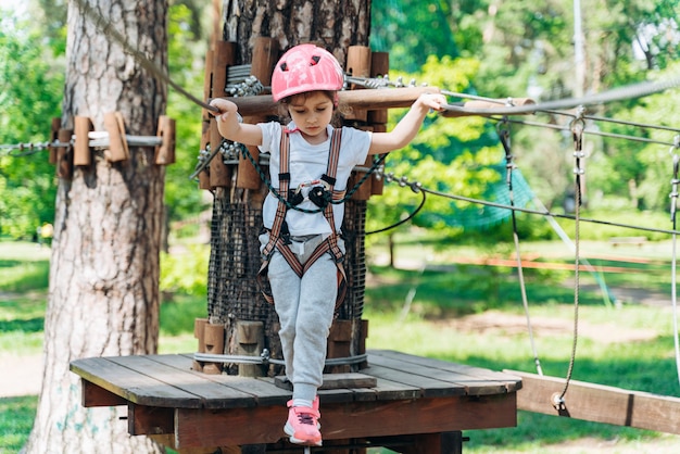 Little girl is standing on a rope, holding a rope with his hands. A child in a rope park passes obstacles