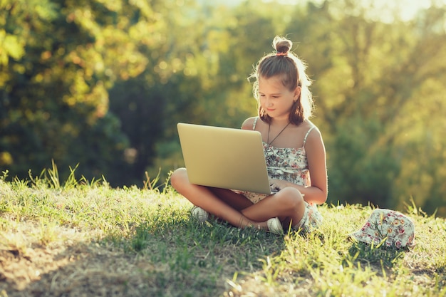 Little girl is talking on a laptop while sitting on the grass in the sun. Dressed in a sarafan and hat