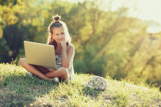 Little girl is talking on a laptop while sitting on the grass in the sun. Dressed in a sarafan and hat