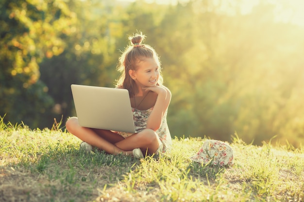 Little girl is talking on a laptop while sitting on the grass in the sun. Dressed in a sarafan and hat