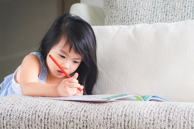 Little girl is writing the book with red pencil on the sofa.