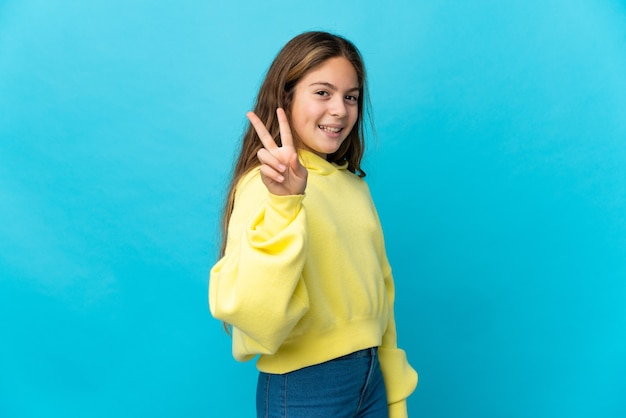 Little girl over isolated blue wall smiling and showing victory sign