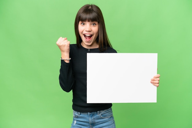 Little girl over isolated chroma key background holding an empty placard and celebrating a victory