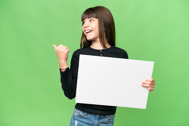 Little girl over isolated chroma key background holding an empty placard and pointing side