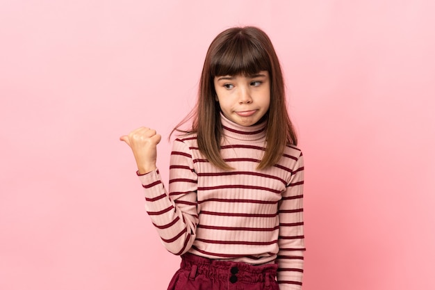 Little girl isolated on pink background unhappy and pointing to the side
