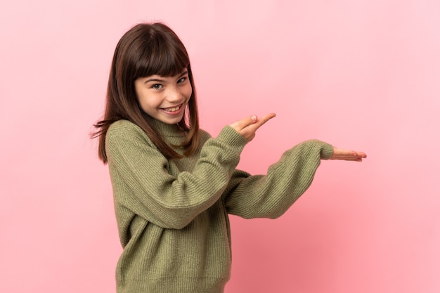 Little girl isolated on pink wall holding copyspace imaginary on the palm to insert an ad