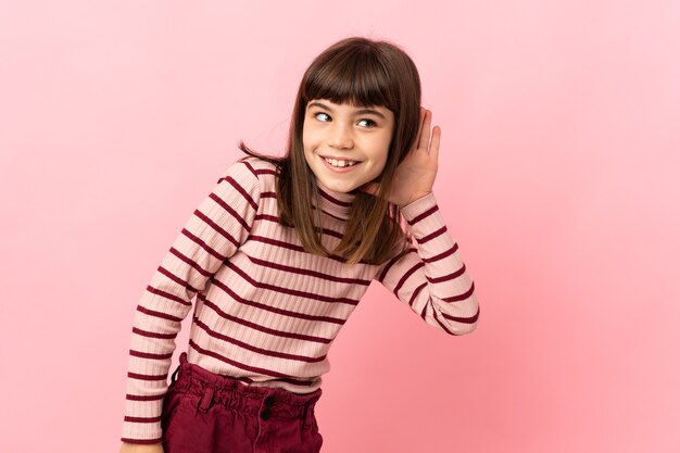 Little girl isolated on pink wall listening to something by putting hand on the ear