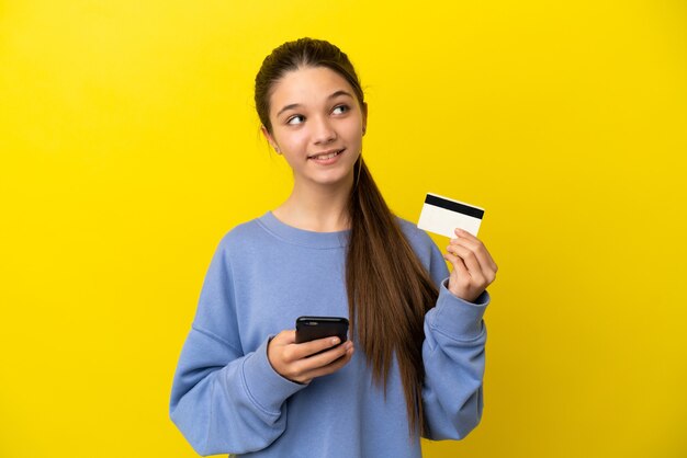 Little girl over isolated yellow background buying with the mobile with a credit card while thinking