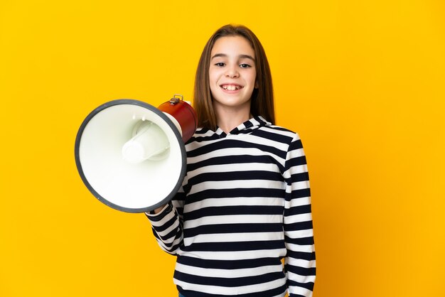 Little girl isolated on yellow background holding a megaphone and smiling a lot