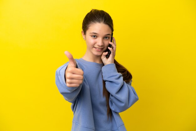 Little girl over isolated yellow background keeping a conversation with the mobile while doing thumbs up