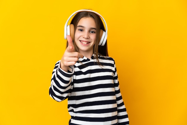Little girl isolated on yellow background listening music and pointing to the front