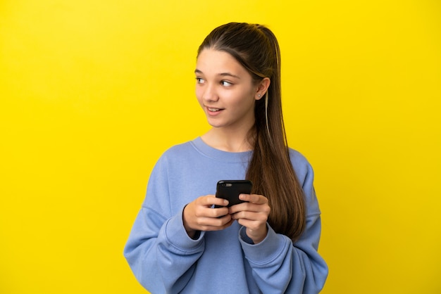 Little girl over isolated yellow background using mobile phone and looking up