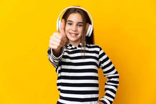 Little girl isolated on yellow wall listening music and with thumb up