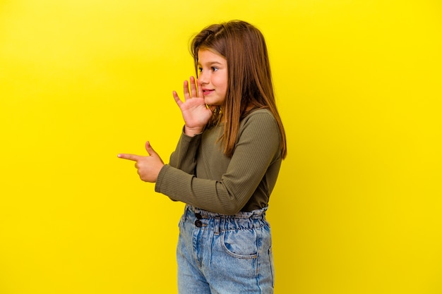 Little girl isolated on yellow wall saying a gossip, pointing to side reporting something