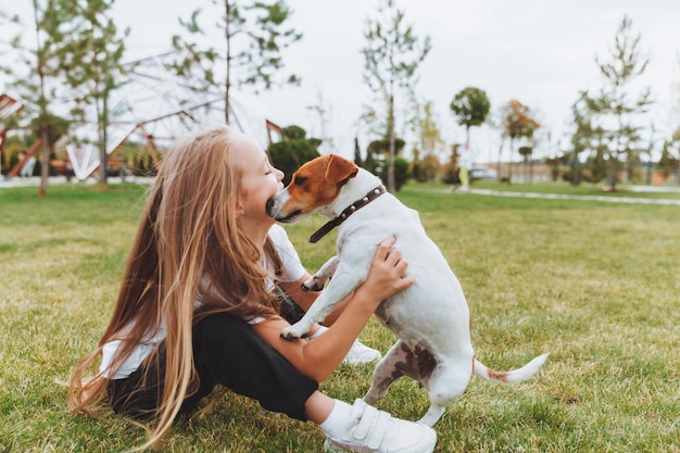 Маленькая девочка целует и обнимает своего джек-рассел-терьера в парке Любовь между хозяином и собакой ребенок держит собаку на руках
