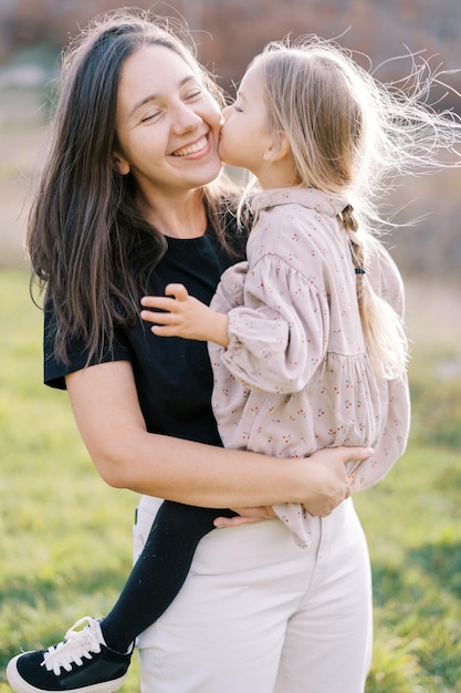 Маленькая девочка целует улыбающуюся мать в щеку, сидя у нее на руках