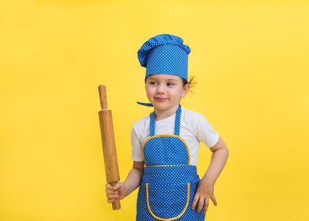 Photo a little girl in a kitchen apron and hat with a rolling pin on a yellow space. looking off to the side.