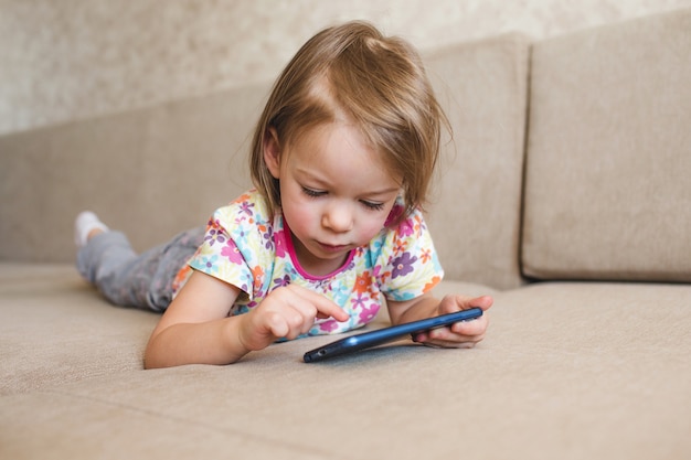 Una bambina è sdraiata sul divano e guarda il telefono