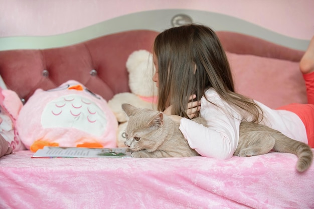 a little girl lies on her bed and reads a book, next to her is a domestic red cat