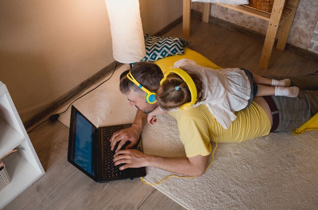 A little girl lies on her father's back and listens to music with yellow headphones while looking at her laptop. the concept of family happiness