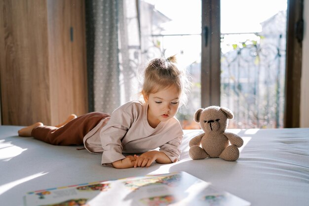 小さな女の子がテディベアと一緒にベッドに横たわって絵本を見ています