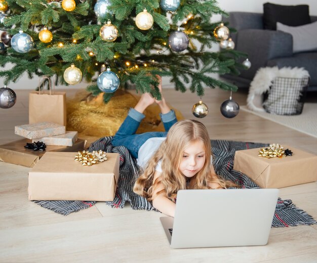 Little girl lies near the Christmas tree with a laptop.  Christmas tree in a large living room.