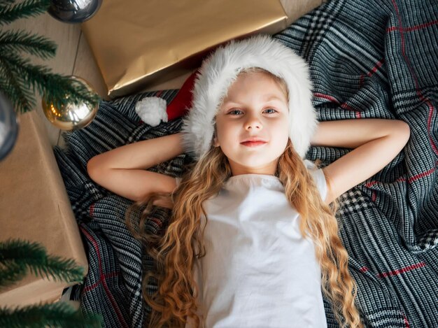 The little girl lies under the tree and dreams. Christmas tree in the large living room.