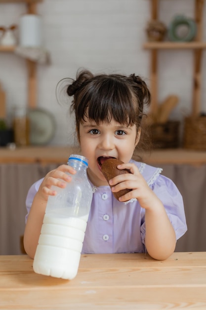 薄紫色のドレスを着た少女は牛乳を飲み、台所のテーブルでパンを食べます。テキスト、バナー用のスペース