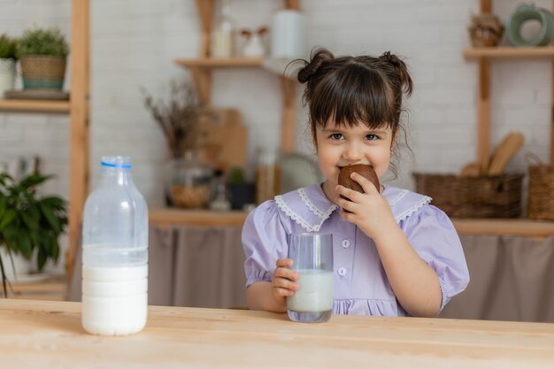 薄紫色のドレスを着た少女は牛乳を飲み、台所のテーブルでパンを食べます。テキスト、バナー用のスペース