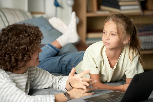 Photo little girl listening to her mom while she explaining her something they lying on bed and using lapt