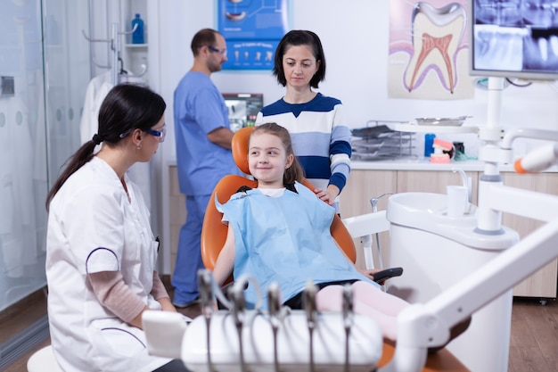 Little girl looking happy at dentist after professional tooth cavity treatment in dental office. Child with her mother during teeth check up with stomatolog sitting on chair.