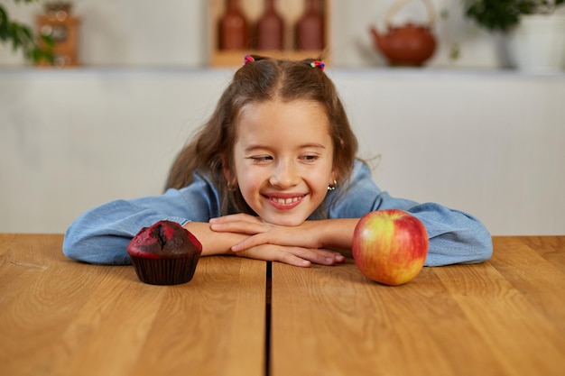 Little girl looking on sweet cake and fresh red apple and choosing in kitchen at home