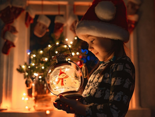 Little girl looks at a christmas lantern illuminating the face in santas hat at night in the room