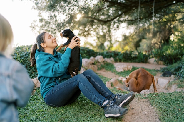 Photo little girl looks at her mother raising a black puppy in her arms while sitting on the grass